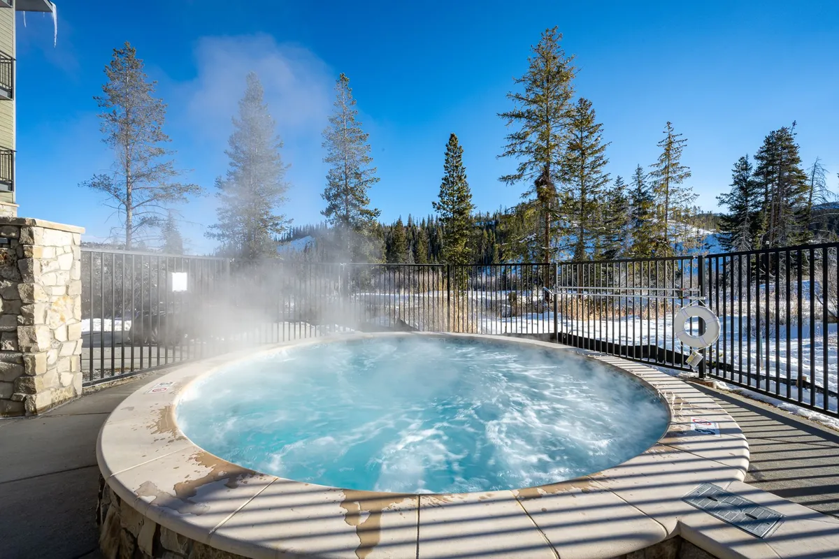 An exterior view featuring a tranquil community hot tub nestled among tall trees, offering a sweeping mountain view in the background. Guests are soaking in the hot tub, relaxing together, and enjoying the peaceful outdoor setting.