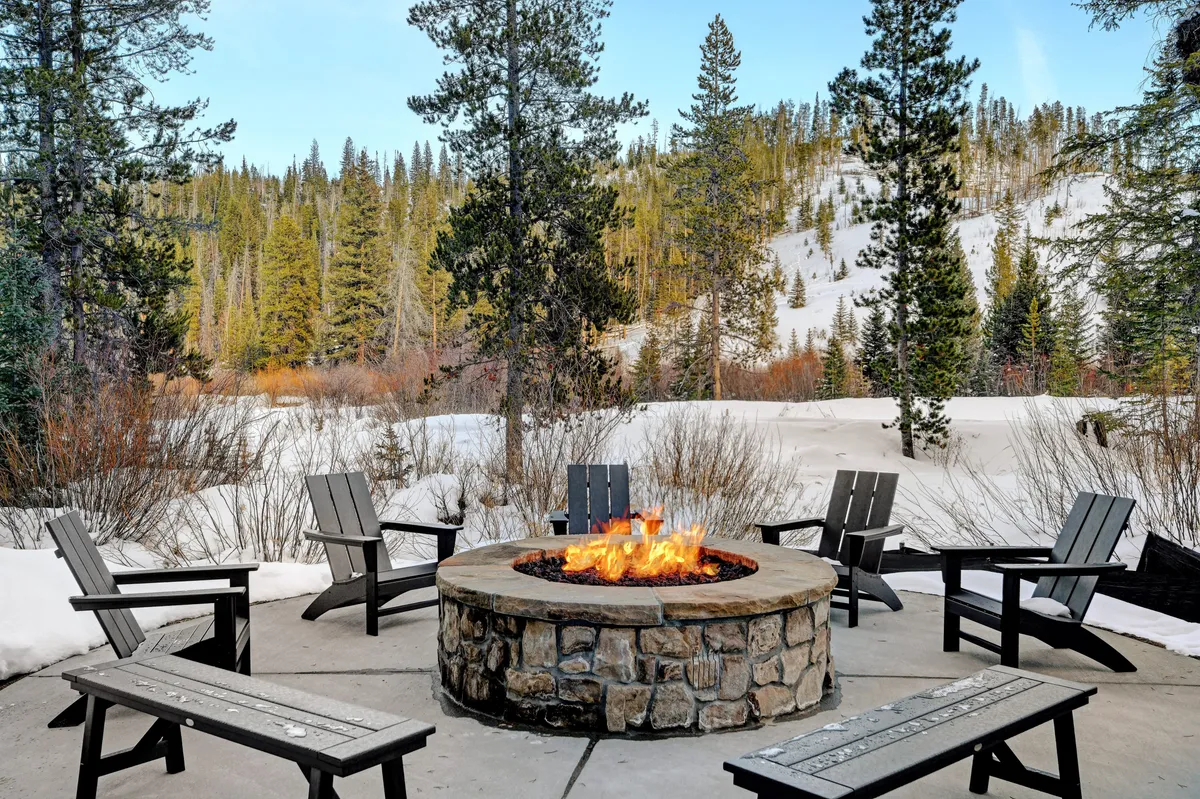 A cozy exterior scene showing a fire pit surrounded by comfortable seating, all set against a snowy landscape. People are gathered around, sharing stories, keeping warm, and enjoying the communal atmosphere under the night sky.