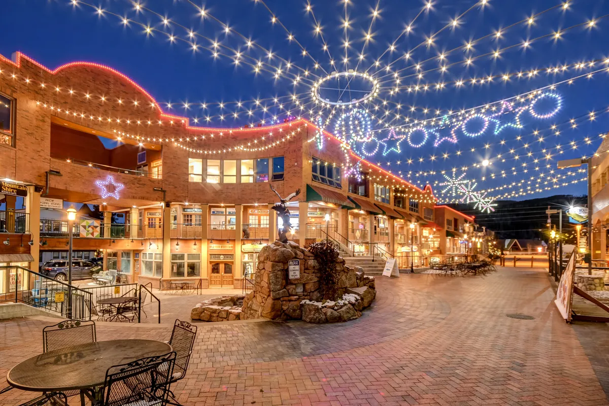 A vibrant downtown square illuminated by string lights, with a central fountain surrounded by shops and restaurants. The scene is lively with people strolling, dining, and enjoying the festive evening atmosphere.