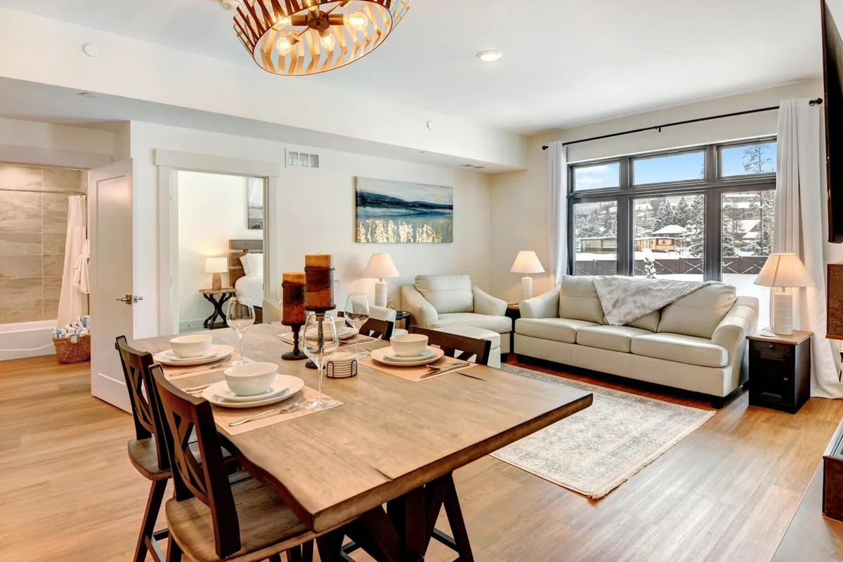 This image centers on a beautifully arranged dining table with place settings, a comfortable sofa, and large windows framing natural views. The scene invites relaxation and togetherness, with a balance of convivial dining and tranquil lounging.