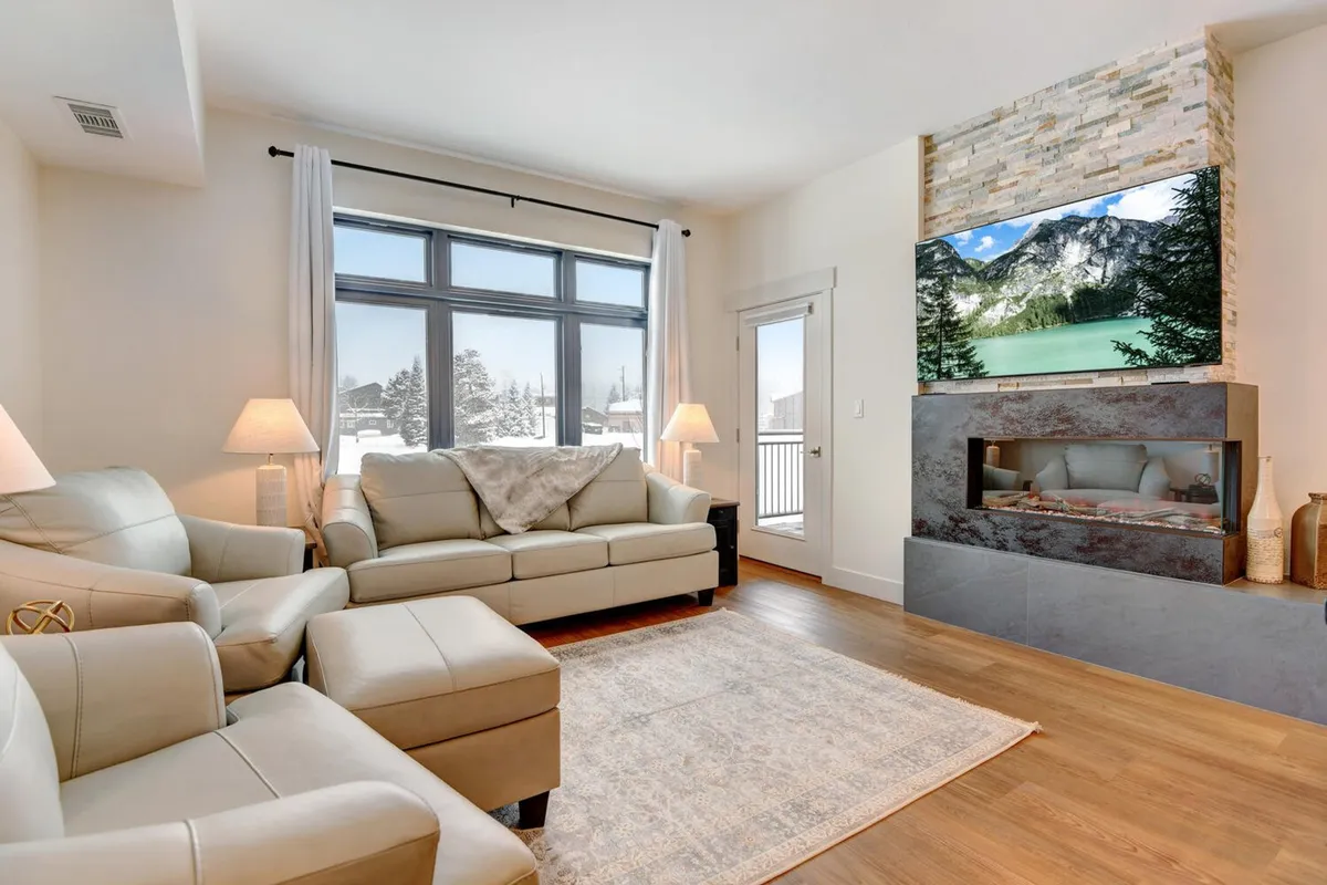 This image showcases a spacious, open living area featuring large windows that frame stunning mountain views, a modern color-changing fireplace, and plush, comfortable seating. The natural light fills the space, creating an inviting, serene atmosphere ideal for both relaxation and entertaining.