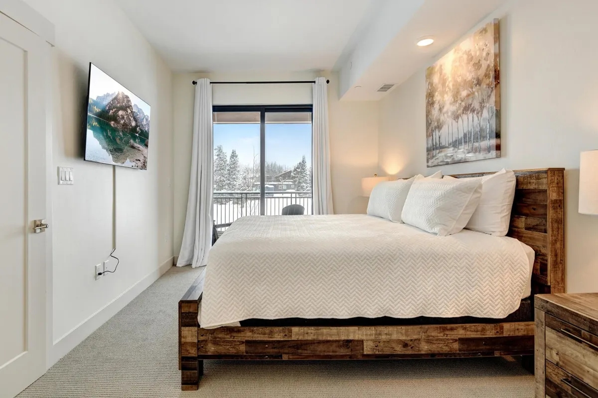 A cozy queen bedroom with a comfortable bed, window offering scenic views, a wall-mounted TV, and tasteful artwork. The space feels inviting, perfect for solo travelers or couples, and provides entertainment and a relaxing environment.