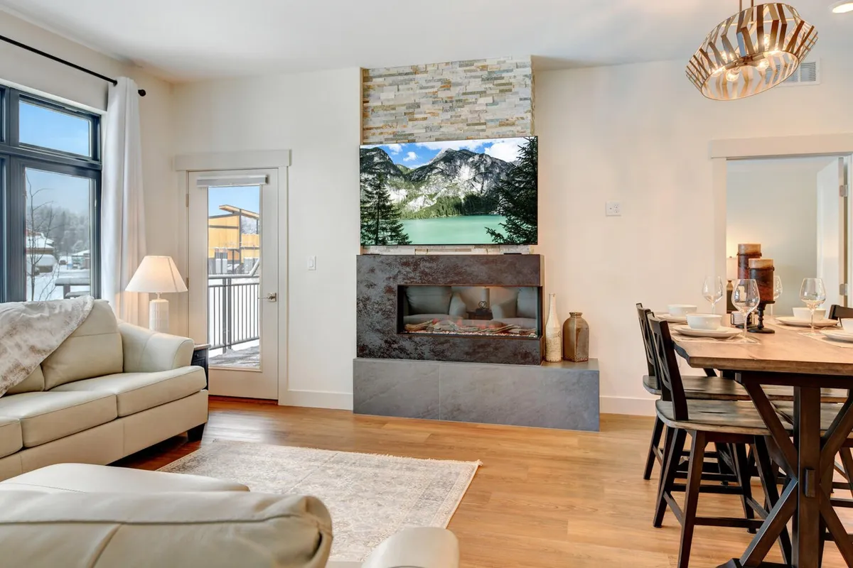 This image showcases a bright, welcoming open-plan living and dining area. Large windows flood the space with natural light, offering glimpses of the surrounding mountain scenery. The modern fireplace adds a luxurious, cozy touch, while the dining table is elegantly set, inviting guests to enjoy a meal together. The scene conveys comfort, warmth, and a sense of upscale relaxation.