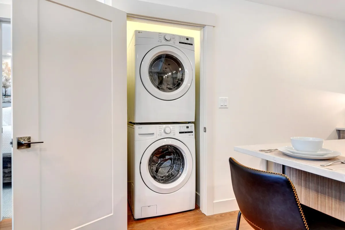 A cozy, functional interior showcases a stacked washer and dryer next to a dining table and chair. The space feels homey and efficient, with laundry appliances seamlessly integrated into the living area.