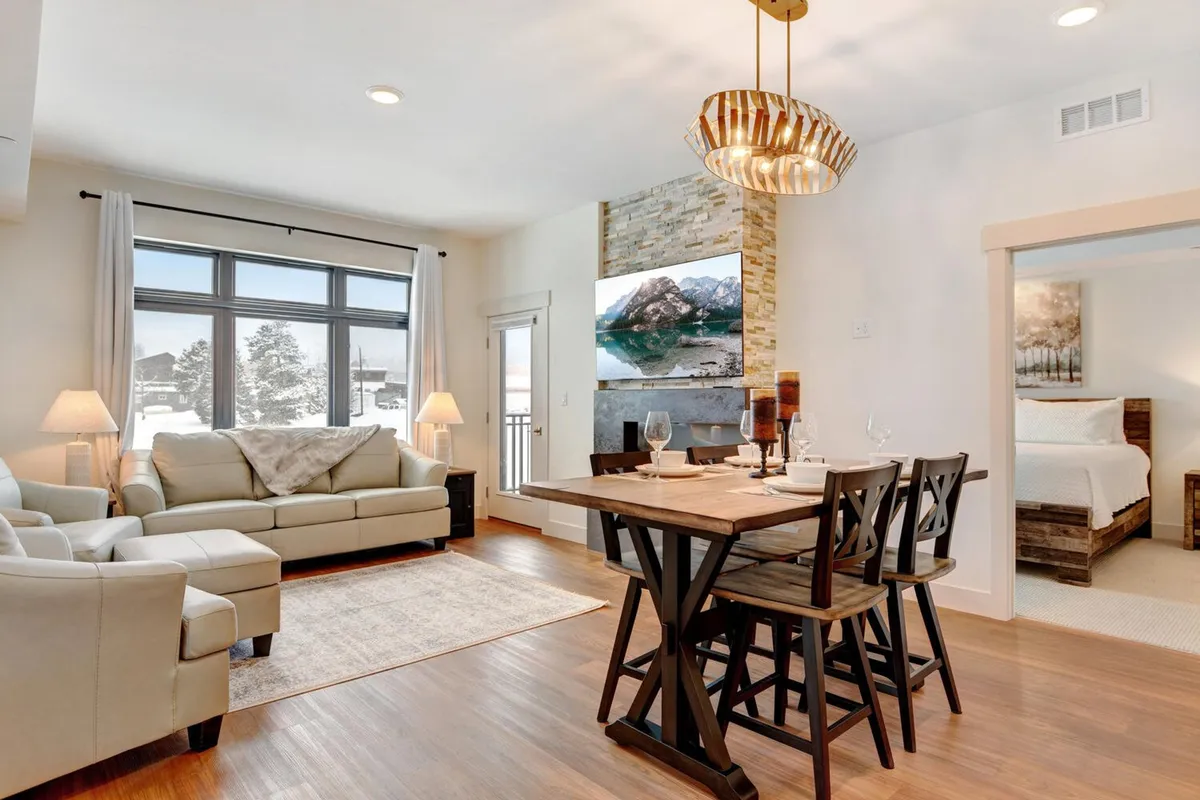The image features a warm, welcoming interior with a plush sofa, chic dining table, modern fireplace, and stylish wall art. The space encourages socializing, dining, and relaxing—showing off the seamless connection between the living and dining areas. The oversized TV is visible, reinforcing entertainment options.
