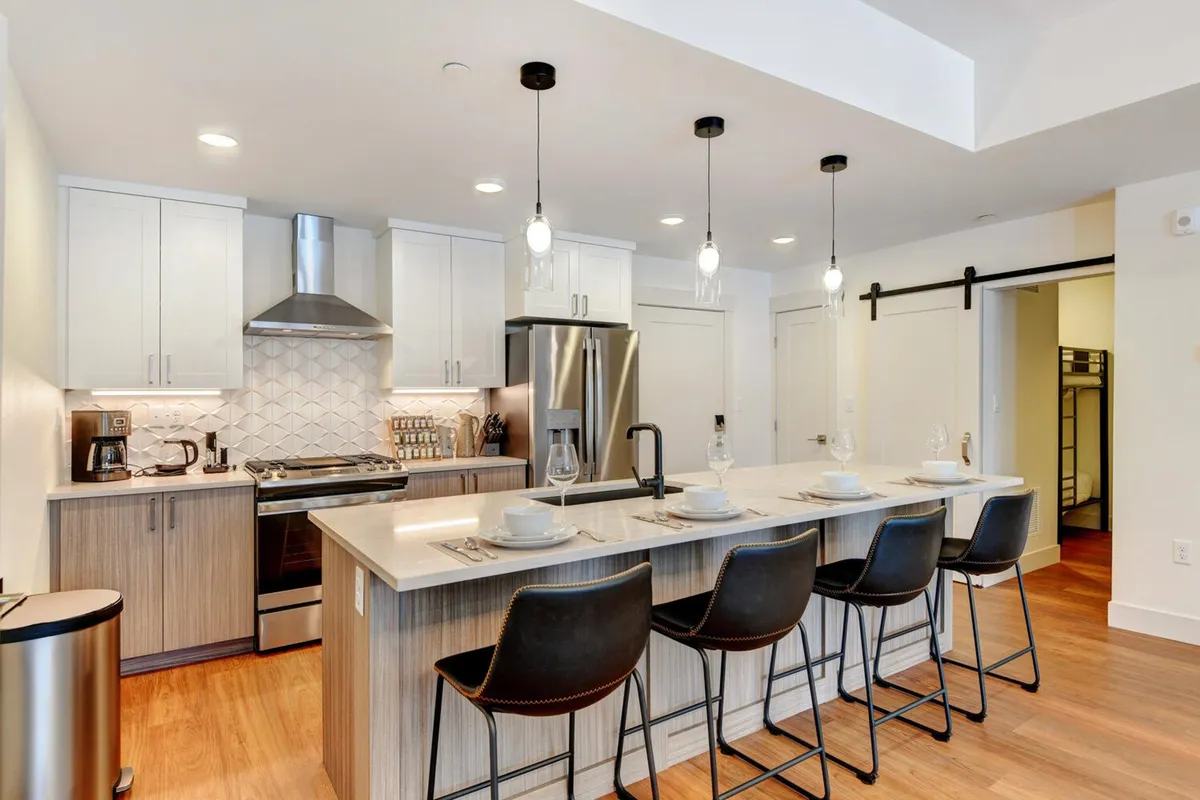 The image showcases a bright, contemporary kitchen with a spacious island, sleek stainless steel appliances, and elegant pendant lights. The setting is designed for both functionality and style, making it perfect for cooking, entertaining, and gathering with friends or family.