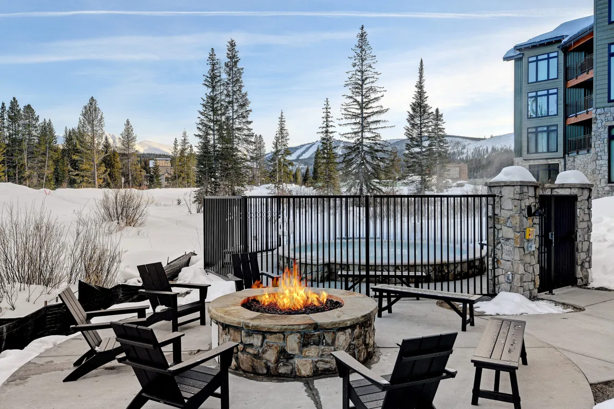 This exterior image features a cozy fire pit area surrounded by seating, set against a snow-covered landscape. In view is a communal hot tub, inviting guests to relax outdoors no matter the season. The scene suggests a perfect spot for families to gather after a day of activities, enjoying both warmth and scenic mountain surroundings.